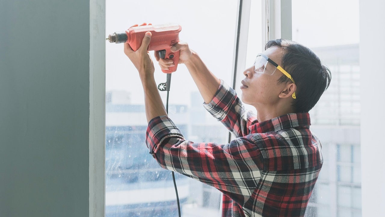 A person wearing safety glasses and rolled-up sleeves using a power drill to repair a window frame.