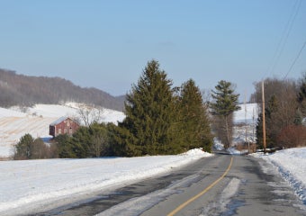 A snowy landscape with a country road passing by a red barn and a stand of evergreen trees